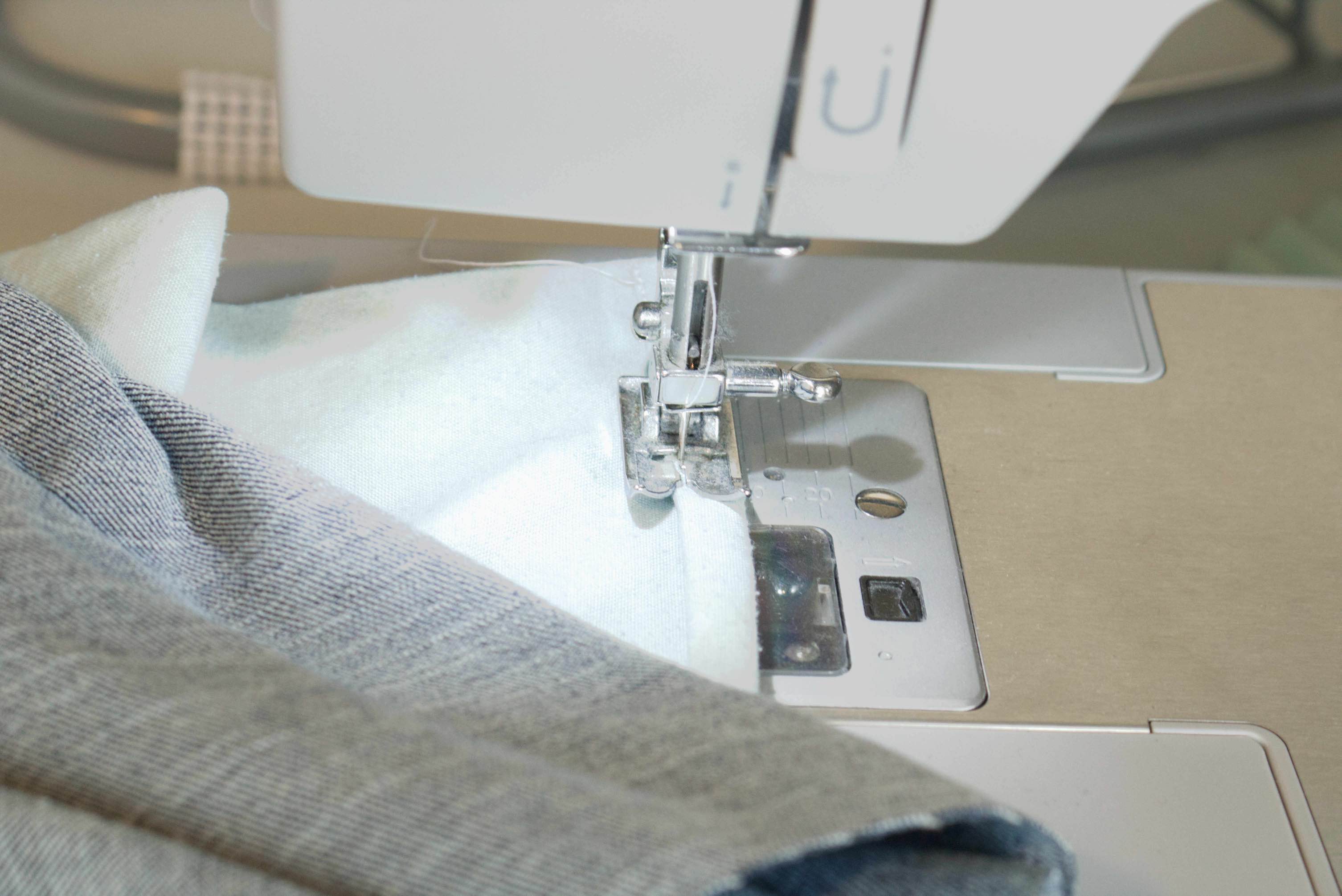 The rolled hem under the sewing machine foot, with the new bottom of the pocket at the edge of the foot, and the needle sunk into the opposite edge of the rolled hem.