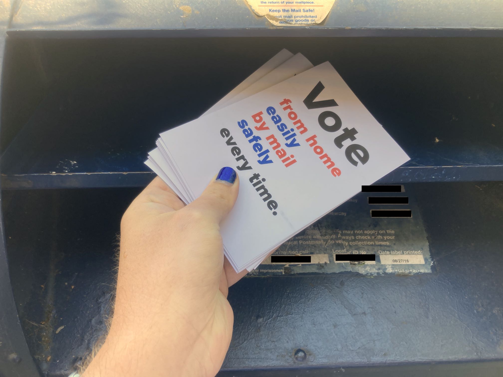 My hand inserting some postcards into a blue USPS mailbox.