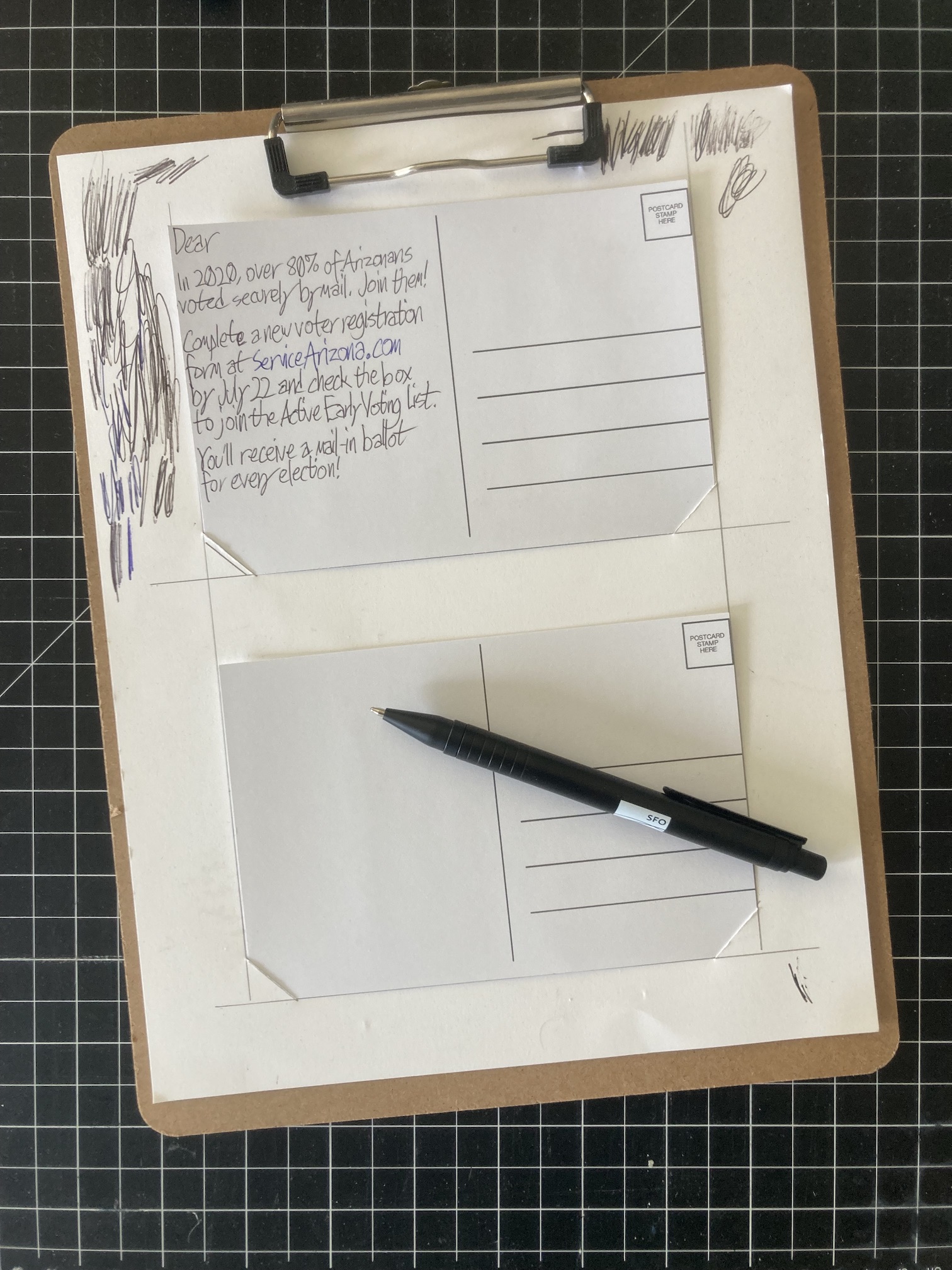 A clipboard holding one postcard holder sheet, with a postcard in each space. Each postcard is back-side-up. The upper postcard has a script written in its left half, while the lower postcard is blank. The perimeter of the sheet is filled with scribbles made to get dried-up pens flowing again.