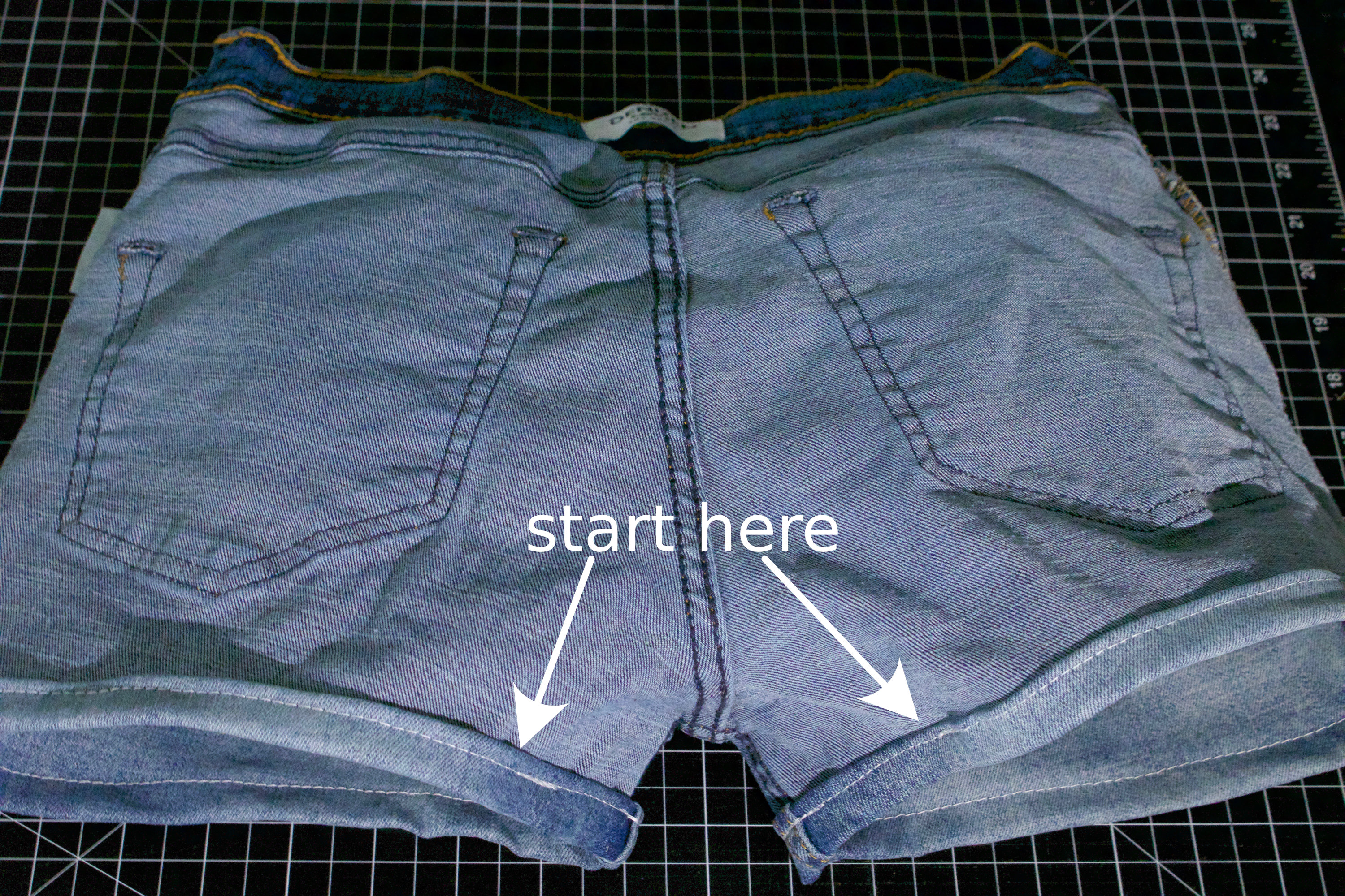 Photo of completed shorts, inside out, rear side up, with a pair of arrows pointing to the aforementioned point on the hems, both originating from the text “start here”.
